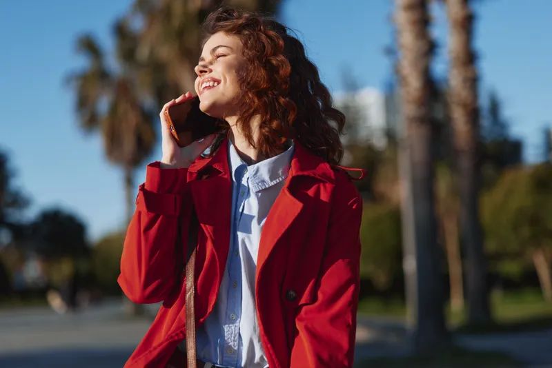 Young woman in a red coat chats on her phone outdoors, sunlit palm trees and a lively street scene create a spontaneous cheerful moment.