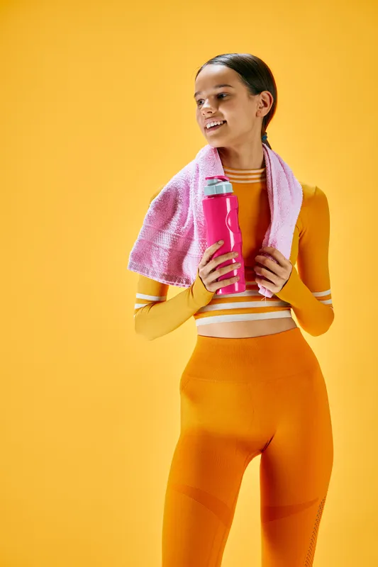 Athletic woman in bright orange workout gear with a pink water bottle and towel around her neck, posing in a vibrant studio for fitness and healthy living.