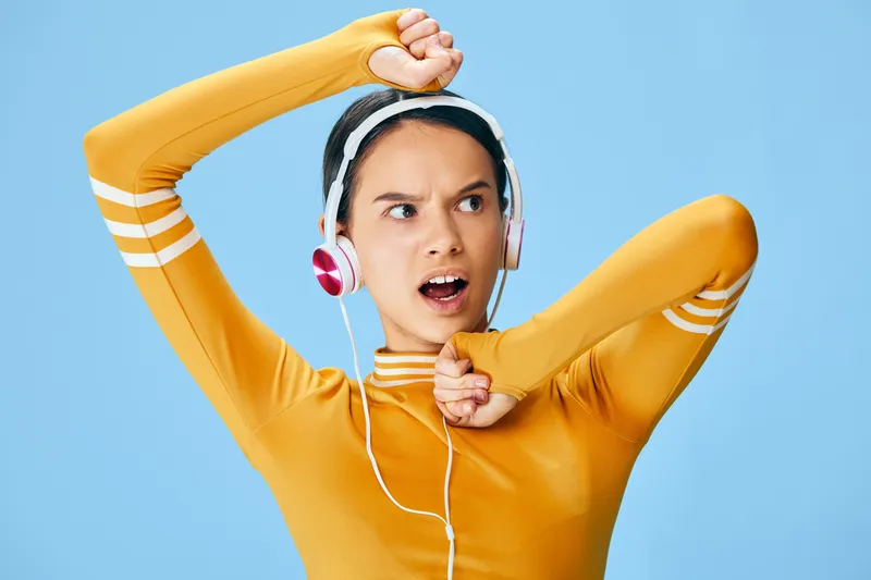 Young fit woman in a yellow long sleeve workout top wearing pink headphones, stretching arms in a dynamic fitness pose with visible headphone cable, energetic sports concept on a clean blue studio