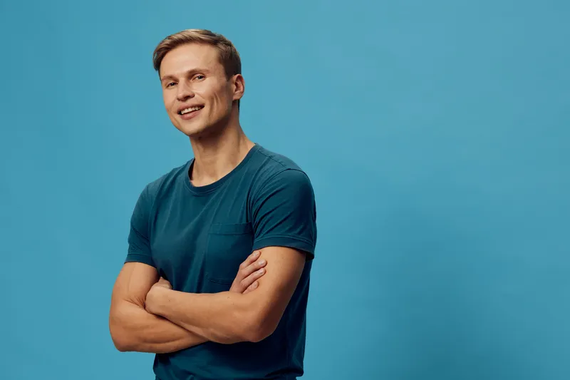 Smiling man young adult with short brown hair wearing casual blue t-shirt standing with arms crossed against a vibrant blue background studio portrait cheerful face.