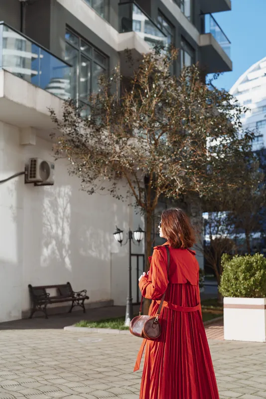 A stylish woman in a crimson maxi dress walks down a sunlit courtyard, her back to the camera, with modern buildings framing the scene and a leather bag swinging.