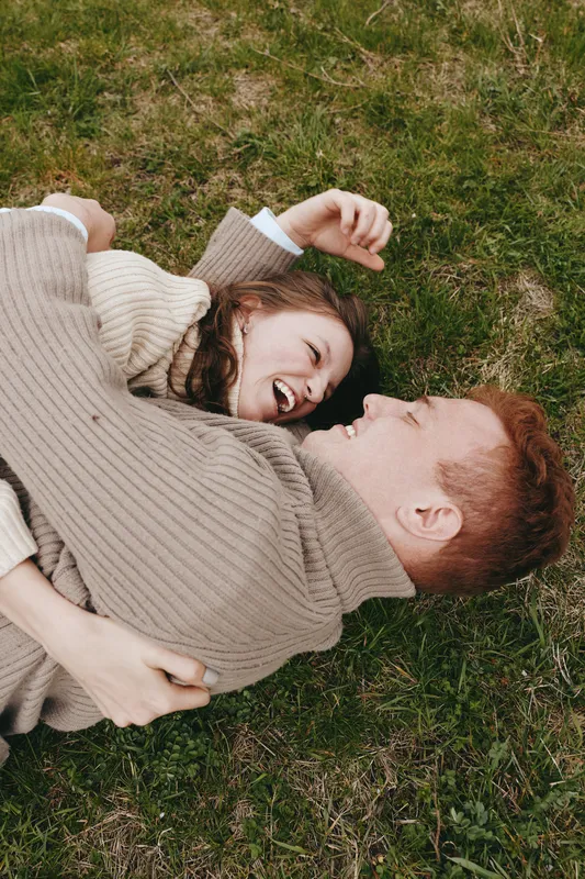 Cheerful couple outdoors, lying on grass, laughing together in cozy sweaters, sunlit moment, relaxed pose, natural colors, light breeze, intimate connection, carefree mood