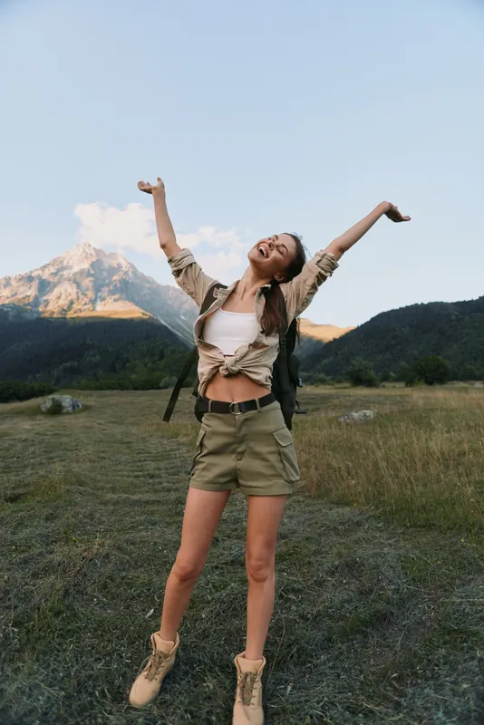 Young woman stands in a mountain meadow with arms raised in celebration wearing khaki shorts and a tied shirt, exuding freedom, energy and outdoor joy.