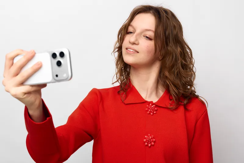 Young woman with curly hair wearing red sweater takes selfie with smartphone on white background. Concept of social media, technology, and casual lifestyle portrait.