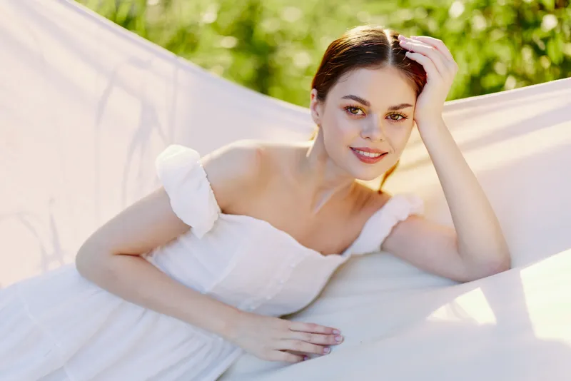 Beautiful young model in a white dress relaxing outdoors, exuding a serene emotion in a lush green setting ideal for seasonal themes