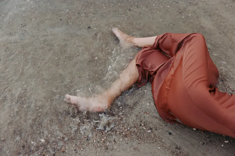 A person lies on a wet, sandy surface with legs extended and feet bare, wearing red-orange pants. The scene conveys exhaustion, vulnerability, and a moment of stillness.