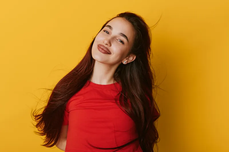 Young woman with long hair wearing red shirt smiling and posing against yellow background, beauty portrait with confident expression and casual style in studio lighting.