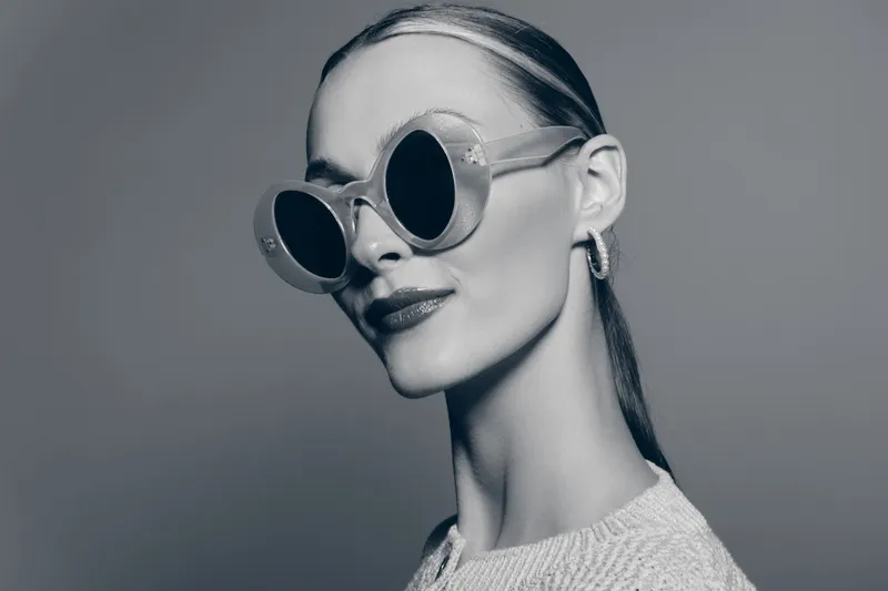 Stylish white woman with fair skin wearing round sunglasses and hoop earrings poses confidently in studio. Black and white portrait with modern fashion and glamour concept.