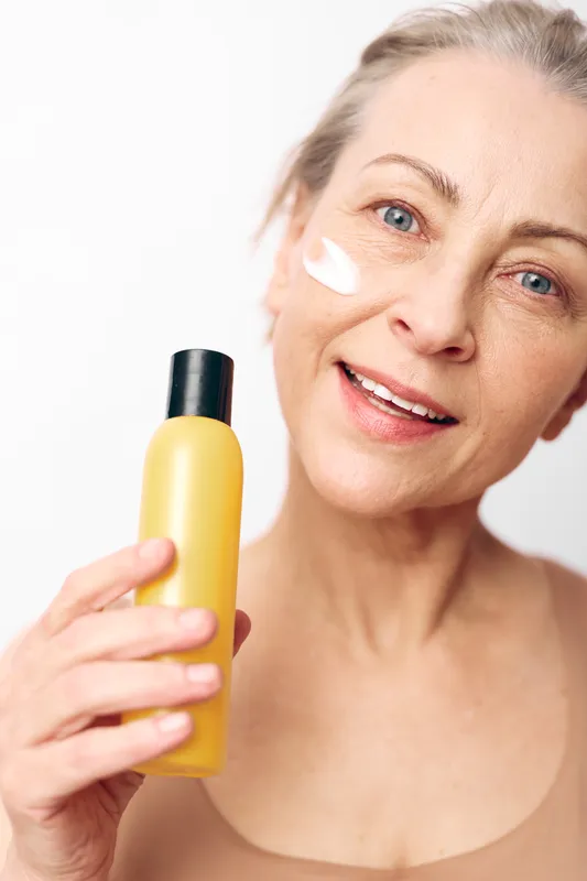 Smiling middle aged woman with clear skin applies white cream on cheek holding yellow bottle. Concept of skincare, natural beauty, moisturizing, and self care routine for healthy skin.