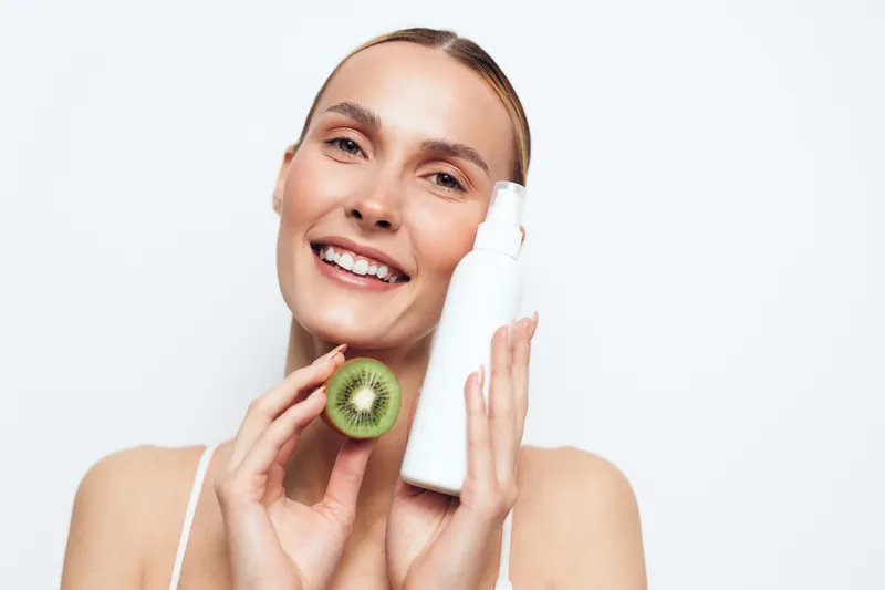 Smiling woman holding kiwi fruit and white cosmetic bottle, fresh skincare concept, natural beauty, clean background, healthy lifestyle, radiant face, wellness routine