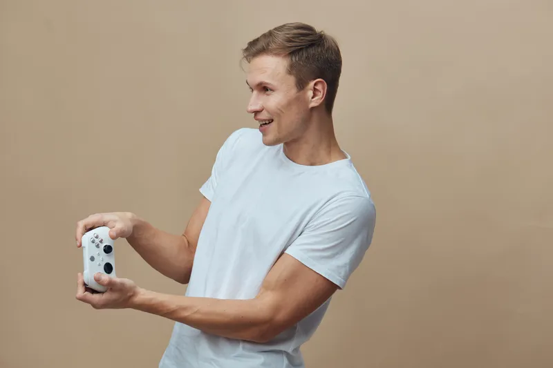 Smiling man holding gaming controller with both hands, wearing casual white t-shirt, looking to the side, in front of plain beige background, studio portrait, lifestyle concept