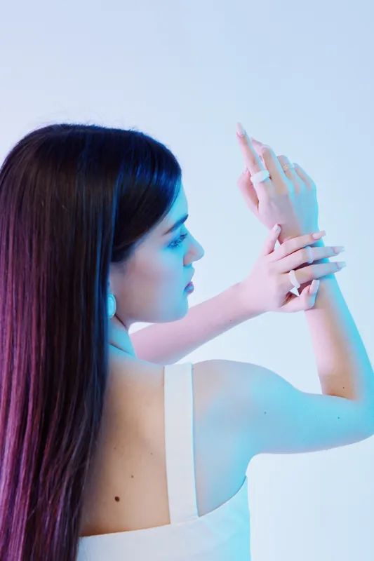 Young woman with long dark hair poses with hands raised, showing elegant rings and earrings. Soft blue lighting highlights her beauty and fashion style in a studio setting.