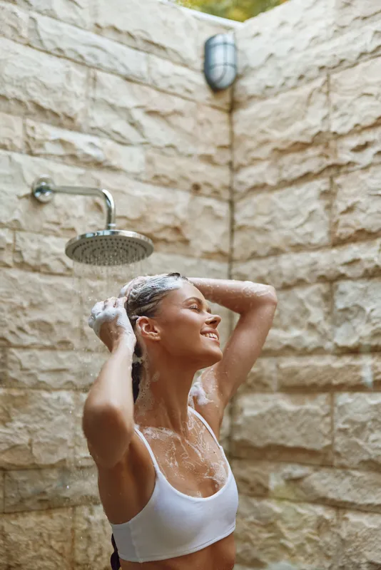young woman washing hair under outdoor shower, relaxed expression, natural light, soap foam, summer wellness, stone wall background