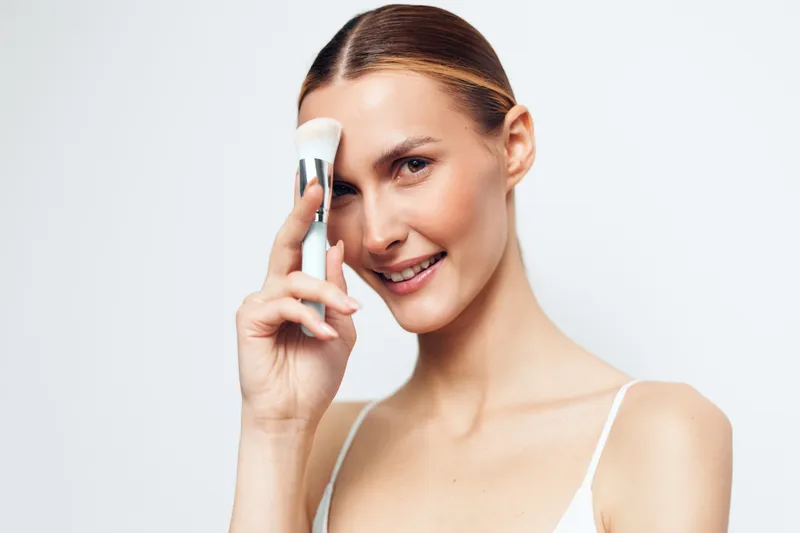 Smiling woman with clear skin applying white face cream with a brush on her forehead. Close-up on a white background. Concept of skincare, beauty, and self-care routine.