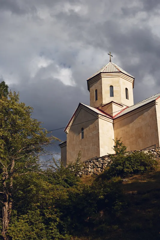 Ancient stone church perched on a hillside under a dramatic cloudy sky, surrounded by trees, showcasing historic architecture, and a tranquil rural landscape with a weathered tower