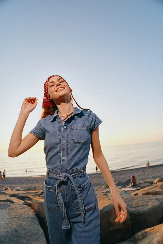 lifestyle woman in modern boho-western denim street style enjoying ocean breeze with warm film color tones during sunset at rocky beach outdoors
