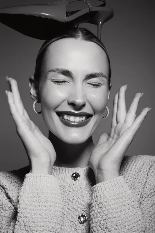 Smiling Caucasian woman with light skin and closed eyes holds hands near face in joyful expression with dark background.