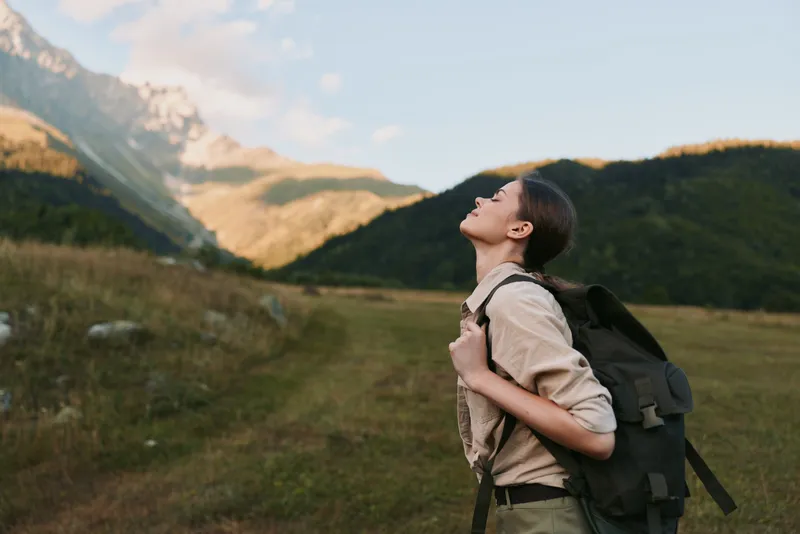 Young woman with a backpack hikes through a grassy valley beneath dramatic mountain scenery, looking upward with a calm, inspired expression in bright daylight.