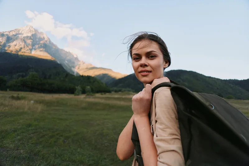 Young woman with a backpack hikes through a grassy meadow with mountains in the background, looking ahead with determination and a calm smile.