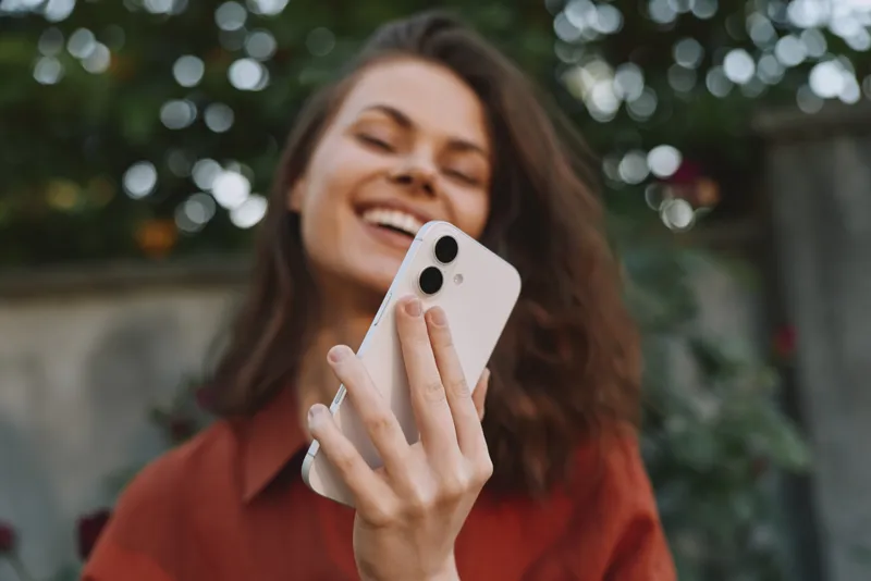 A smiling woman in casual clothes holds a smartphone for a selfie in an outdoor setting with sunshine and greenery, capturing a cheerful lifestyle moment