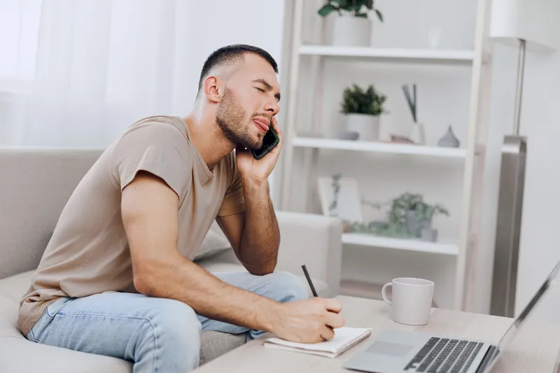 Young man taking notes while on a phone call in a modern, cozy workspace. Bright natural light enhances the relaxed atmosphere, inspiring productivity and creativity.