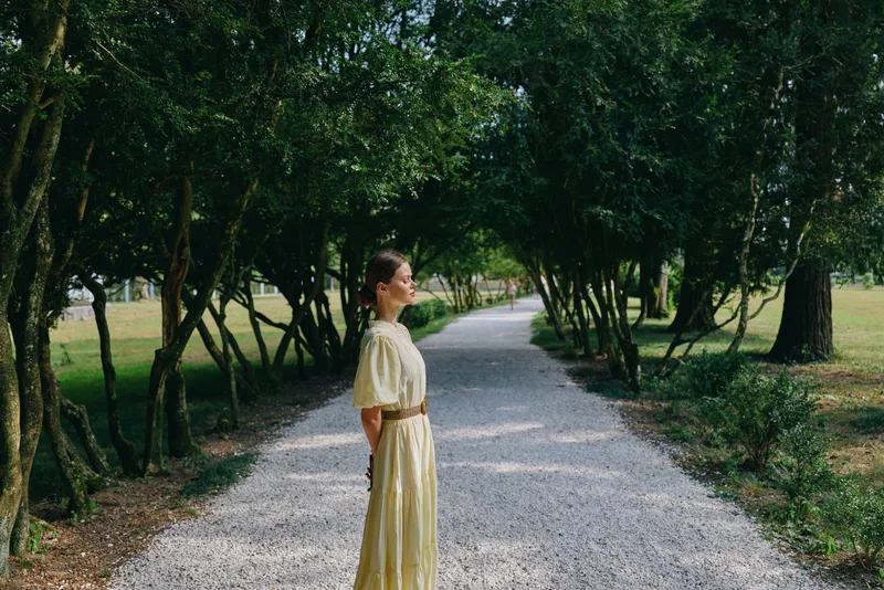 Woman in a dress stands on a gravel path in a park, surrounded by trees and nature, with sunlight filtering through leaves, creating a serene calm mood and outdoor beauty.
