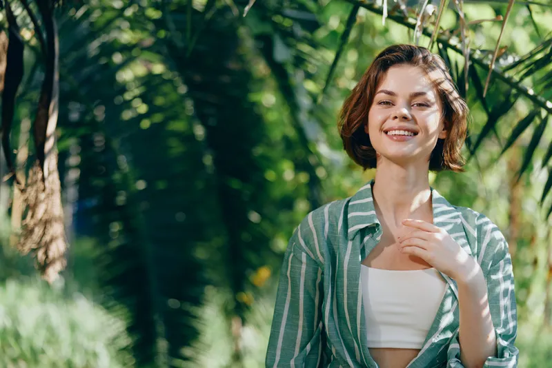 Smiling young woman in a leafy outdoor setting, wearing a striped shirt and light crop top, enjoying warm sunlight and a carefree mood.