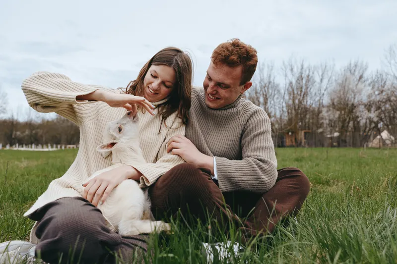 A couple sits on the grass with a playful goat kid, enjoying a sunny outdoor moment. Gentle affection and relaxed lifestyle capture natural happiness on a farm meadow.