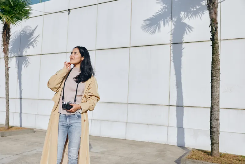 Fashion forward female in a trench coat poses outdoors beside a white wall, holding a camera and looking up with a thoughtful expression, palm shadows frame the scene stock ready