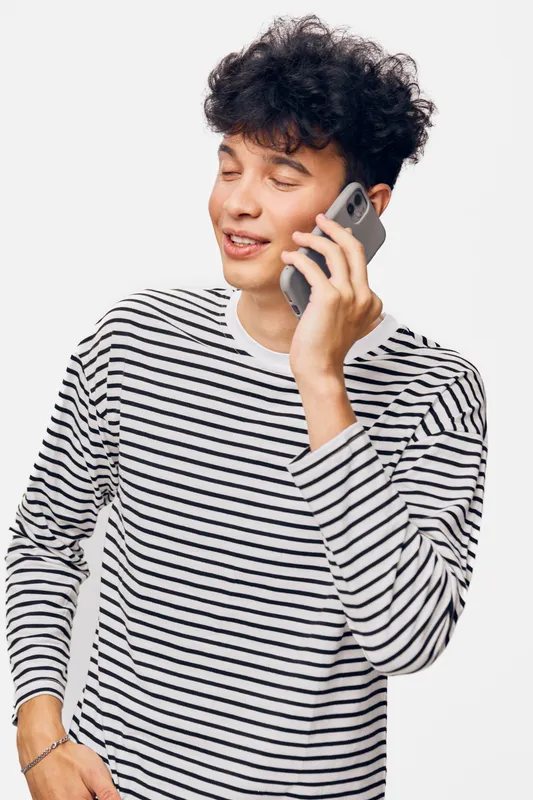 young man talking on smartphone wearing striped long sleeve shirt with curly hair and relaxed expression on white background