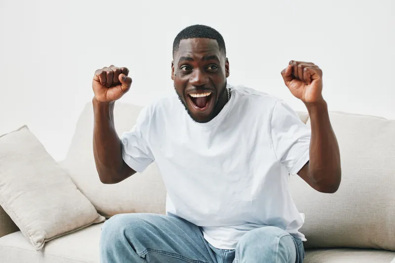 Happy young African man celebrating success in a bright modern living room with a joyful expression, wearing a casual white t shirt and jeans