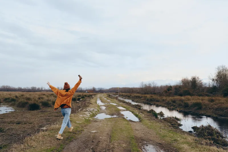 woman outdoors lifestyle, river nature walking, arms raised in happiness, winter countryside trail with wet mud and puddles, cloudy sky, jeans and sweater style, peaceful calm landscape