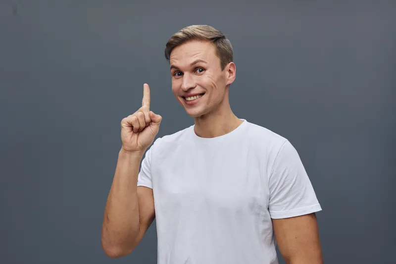 Confident young man with short brown hair wearing casual white t-shirt, smiling and pointing upwards with his index finger, looking directly at camera, on neutral gray background, studio portrait, lif