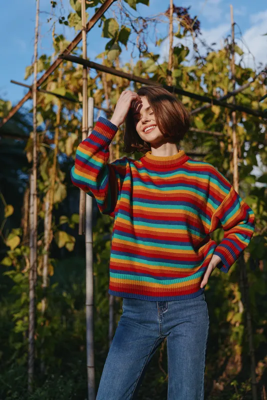 Young woman in a rainbow sweater and casual outfit poses in a garden portrait, smiling woman with short bob and jeans, hand in hair among sunlit vines for relaxed outdoor lifestyle.