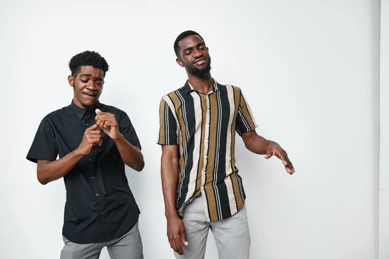 Two young black men with dark skin dancing and enjoying music together in casual clothes on white background with happy expressions and relaxed mood.