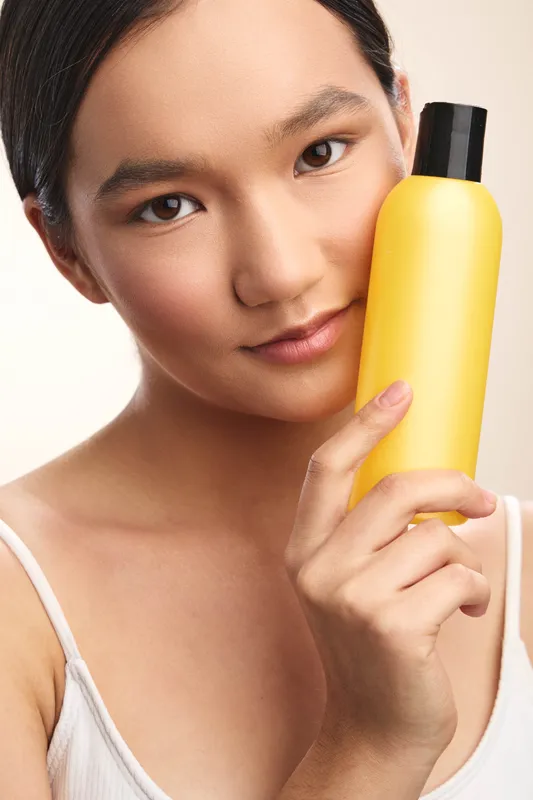 asian woman holds a yellow bottle close to her face in a clean studio, gazing softly at the camera with a subtle smile, presenting a fresh solar glow and modern personal care vibe.