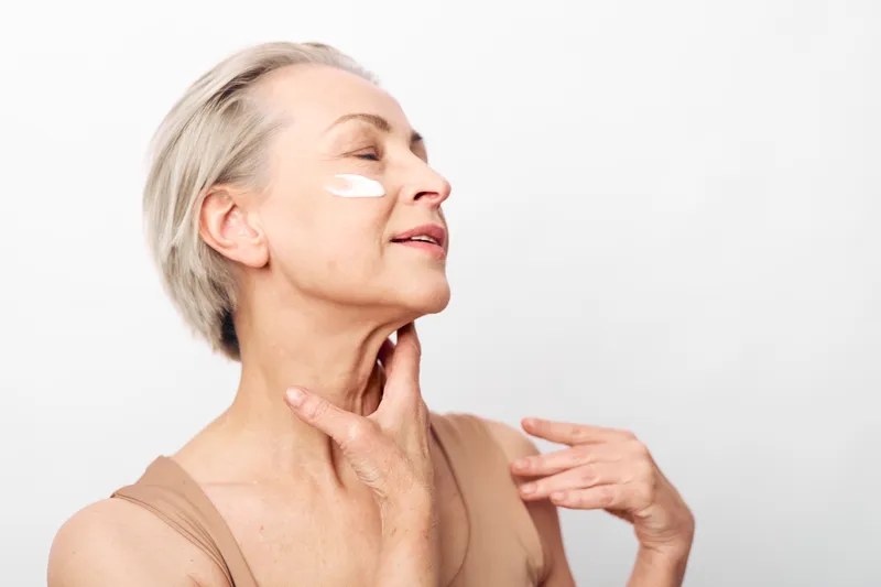 Mature woman with short grey hair applies white face cream on cheek, eyes closed, showing skincare routine and self-care concept on plain white background with soft natural lighting.