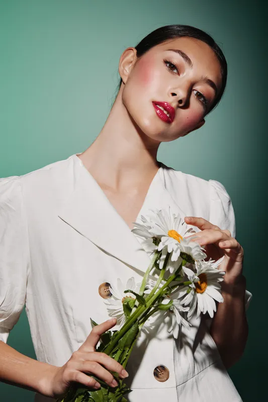Elegant young woman wearing a white outfit holds a bouquet of daisies while posing creatively with glamorous makeup. The light green background enhances the fresh, modern beauty theme.