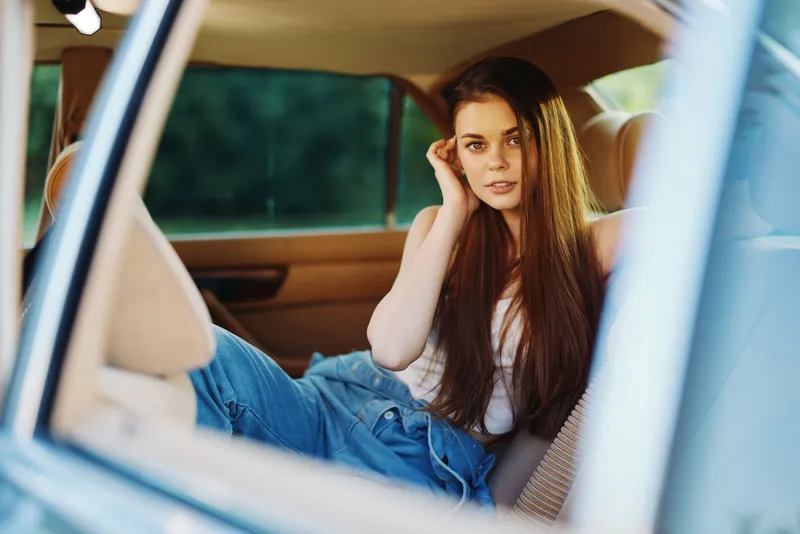 Young woman in casual fashion sitting in a car, showcasing elegance and confidence, with long hair and a relaxed expression against a sunny outdoor background.