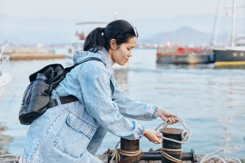 Woman with backpack arranging mooring rope at a harbor, preparing lines for docking, blue jacket, waterfront scene with boats and calm water as activity unfolds