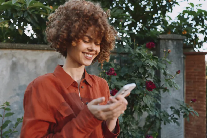 Young woman with curly hair smiles while checking her smartphone in a sunlit garden, conveying casual, everyday connectivity and a relaxed lifestyle moment of modern femininity