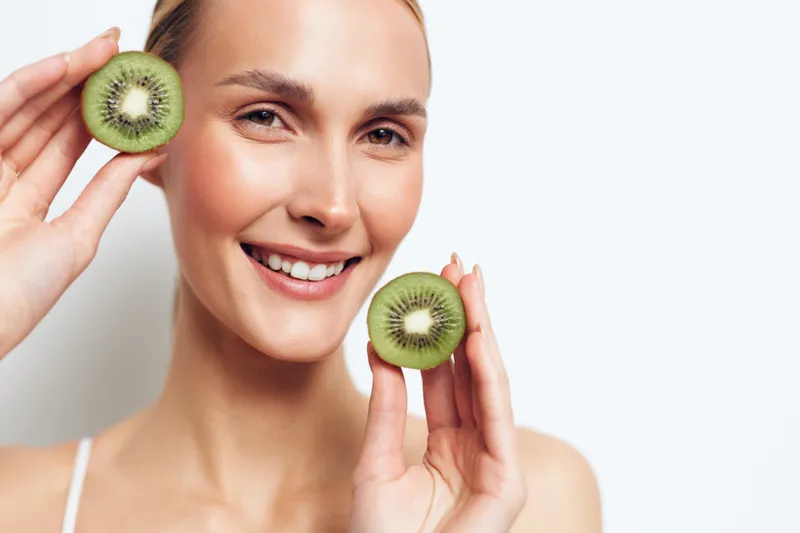 A smiling woman holds two halves of a fresh kiwi fruit near her face against a white background. Concept for healthy lifestyle, natural food, and skincare.