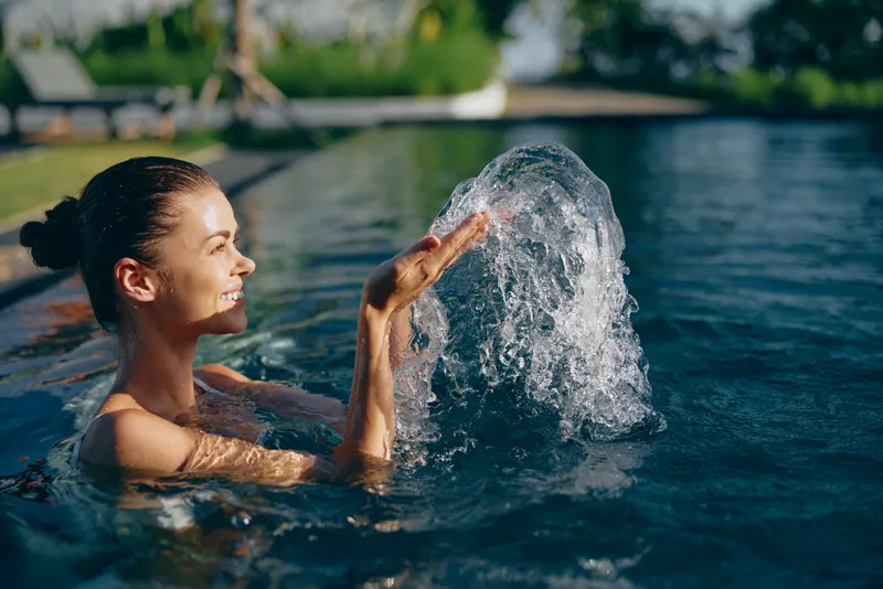 Cheerful woman enjoys a playful splash in a sunlit pool, hands creating water fountain, relaxed outdoor lifestyle moment by the water, summer leisure and joy.