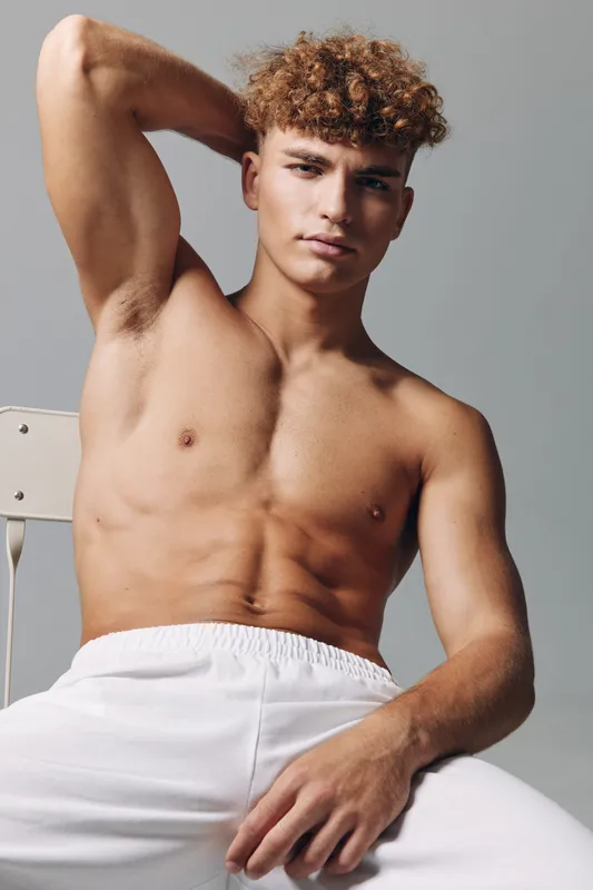 Young man with curly hair posing shirtless in white pants against neutral background, showing muscular physique and confident expression in studio setting.