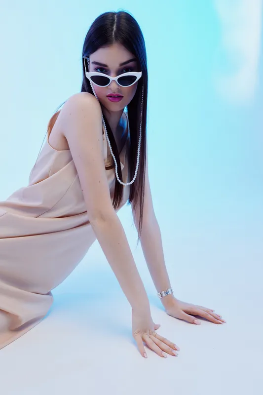 Young woman in beige dress and white sunglasses poses on floor against blue background. Her long hair and pearl necklace emphasize beauty and fashion style.