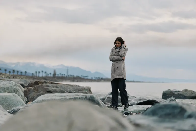 A woman in a gray coat stands alone on rocky shore looking down. Overcast sky and distant mountains create a calm, contemplative mood by the sea. Concept of solitude and nature.