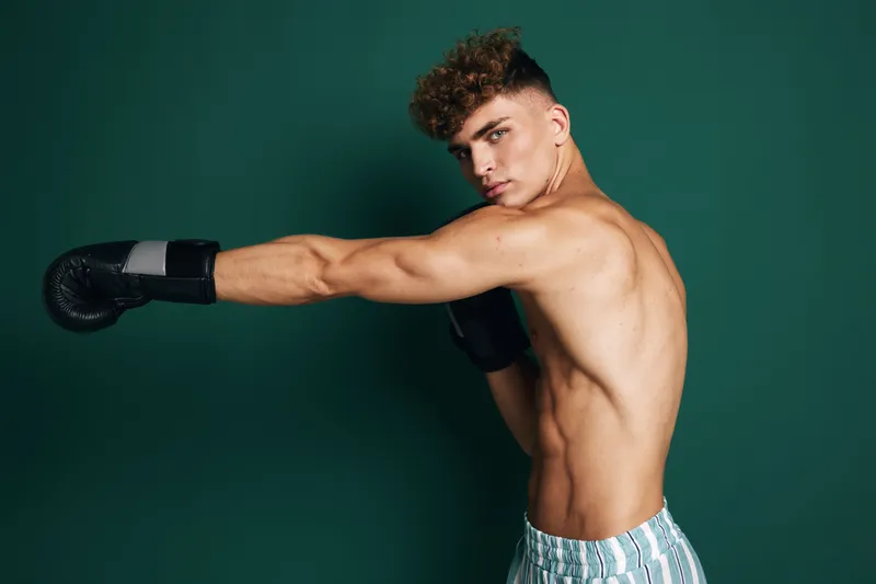 Young male boxer with curly hair wearing gloves and striped shorts throwing a punch against dark green background in studio. Athletic, fit and focused athlete training.