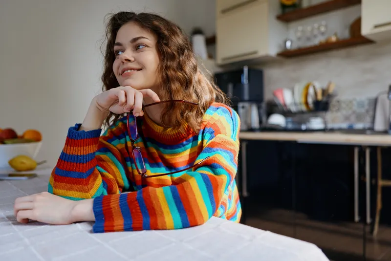 A woman in a bright striped sweater sits at a kitchen table, smiling softly, hands resting under chin. A warm, casual home lifestyle portrait focused on comfort, everyday warmth, and relaxed mood.