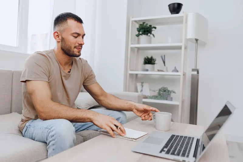Thoughtful man sitting on a couch, enjoying coffee while working on a laptop. A cozy home office atmosphere with a minimalist design, promoting productivity and comfort.
