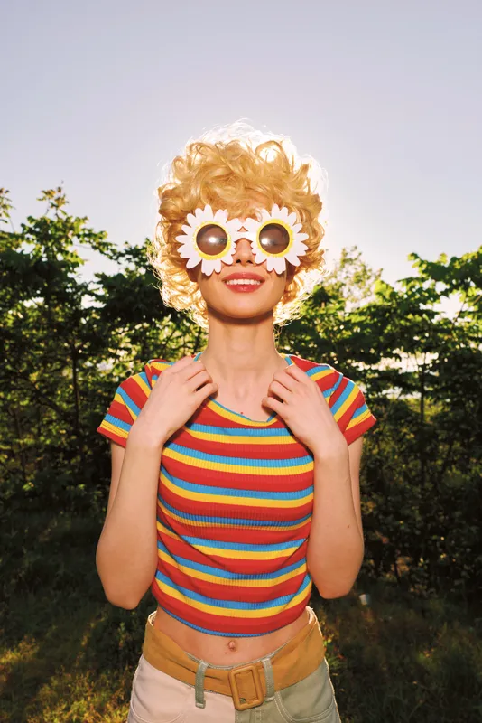 Vibrant woman outdoors, rainbow striped top, playful daisy sunglasses, sunlit moment, candid pose, carefree mood, natural lighting, soft breeze, lush greenery background with glow